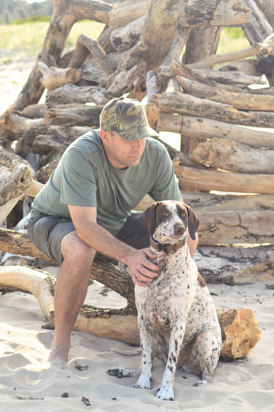 Man sitting on a pile of driftwood with a dog on a sandy beach in Camo Cap
