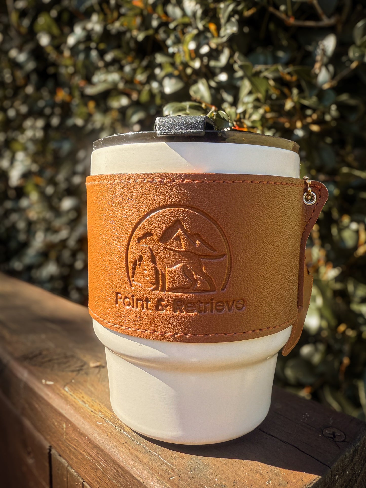 White coffee tumbler with brown leather sleeve featuring a logo and text, placed on a wooden surface with a blurred natural background.