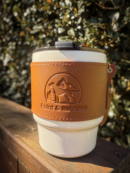 White coffee tumbler with brown leather sleeve featuring a logo and text, placed on a wooden surface with a blurred natural background.