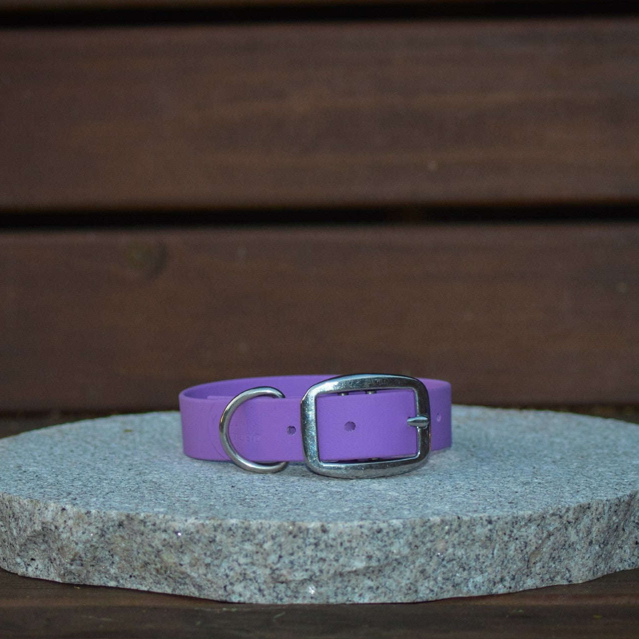A purple leather dog collar with a silver metal buckle, displayed on a round gray stone slab against a dark brown wooden background.
